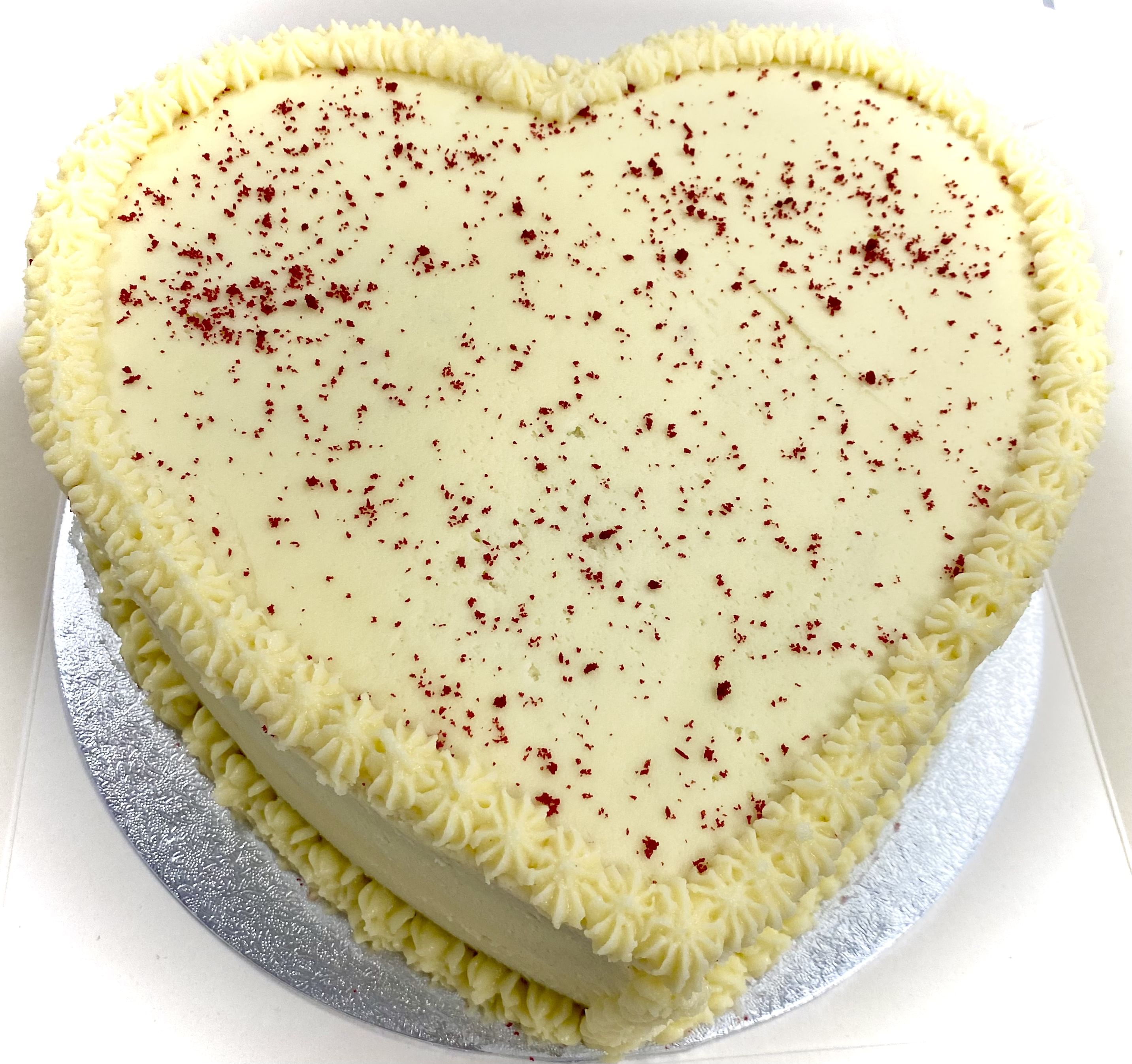A heart-shaped cake covered in white frosting, with a border of piped frosting around the edges. The cake is topped with a light sprinkle of red crumbs and is placed on a silver cake board.